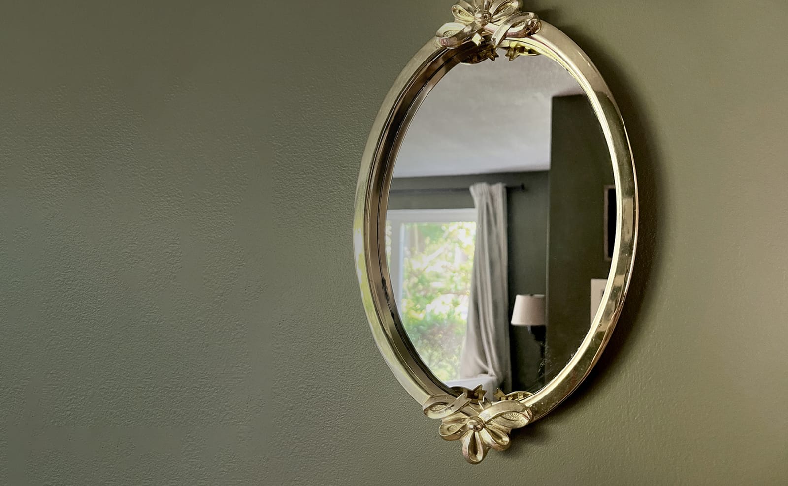 A gilt oval mirror with cast flowers at top and bottom, hung on a wide olive-green wall, reflecting a curtained window and a lamp