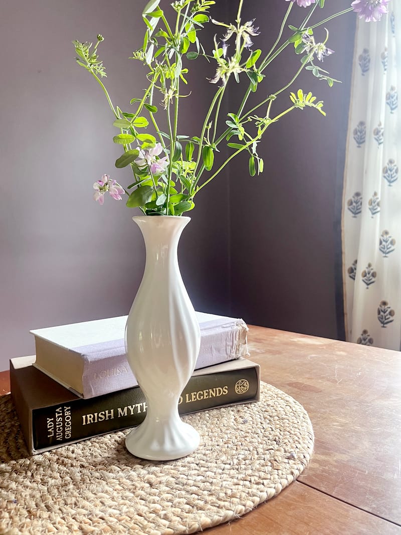 A slender white vase of wildflowers on a stack of books and a jute mat, against a heather wall and block-print curtain