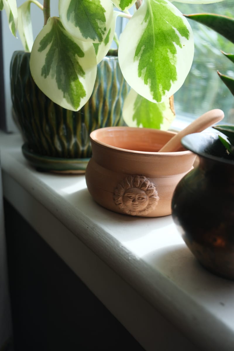 A windowsill with a variegated plant in a green glazed pot, a terracotta bowl cast with a sun face, and a dark metal vessel