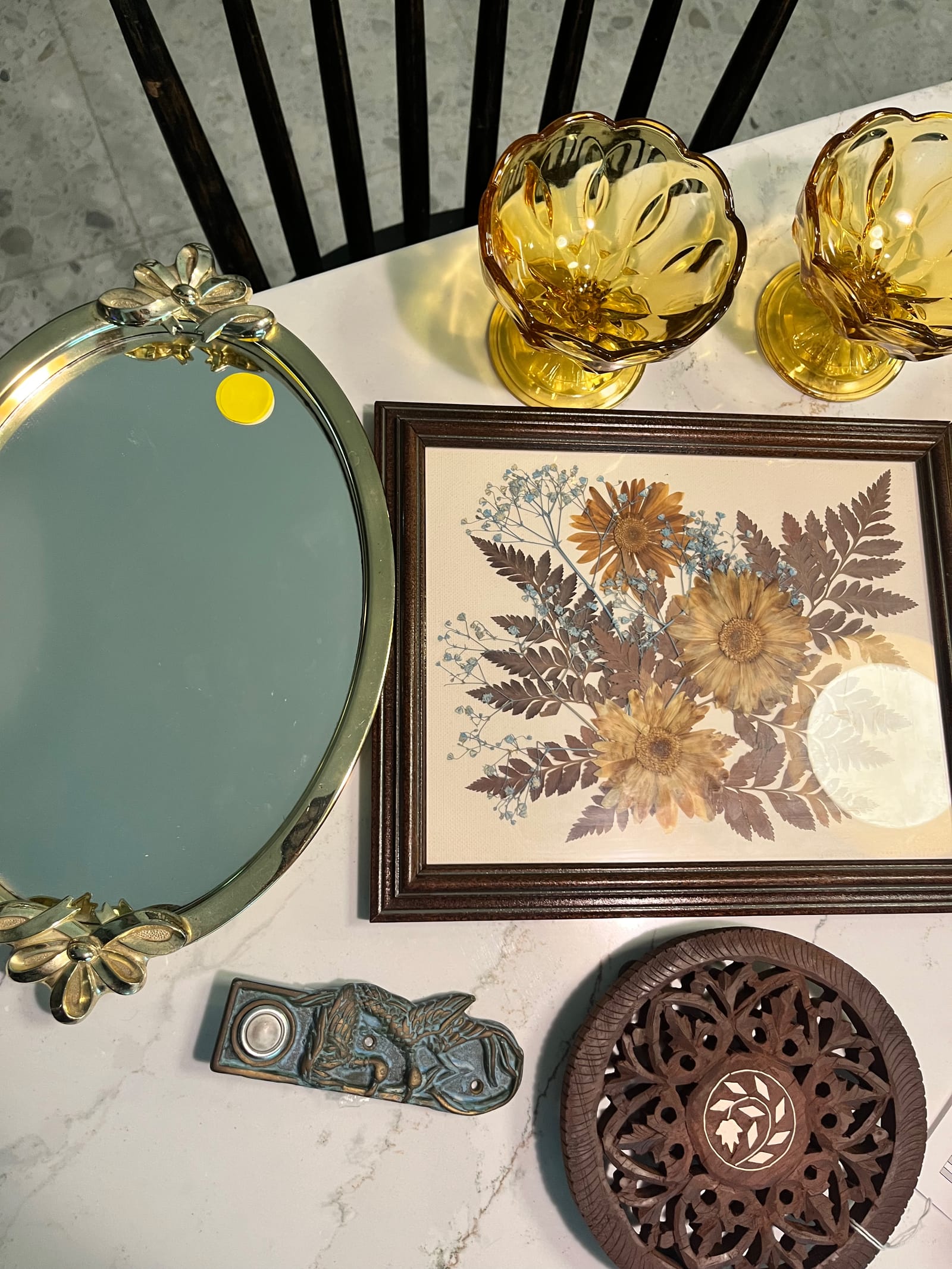 A flatlay of a gilt mirror tray, a framed arrangement of pressed ferns and flowers, amber glass and a carved wooden trivet