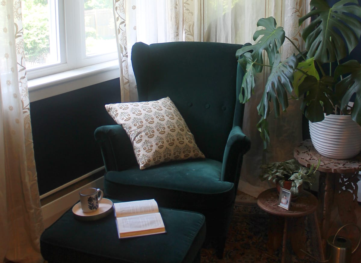 A teal velvet armchair and footstool by a window, with a book, a mug and trailing plants